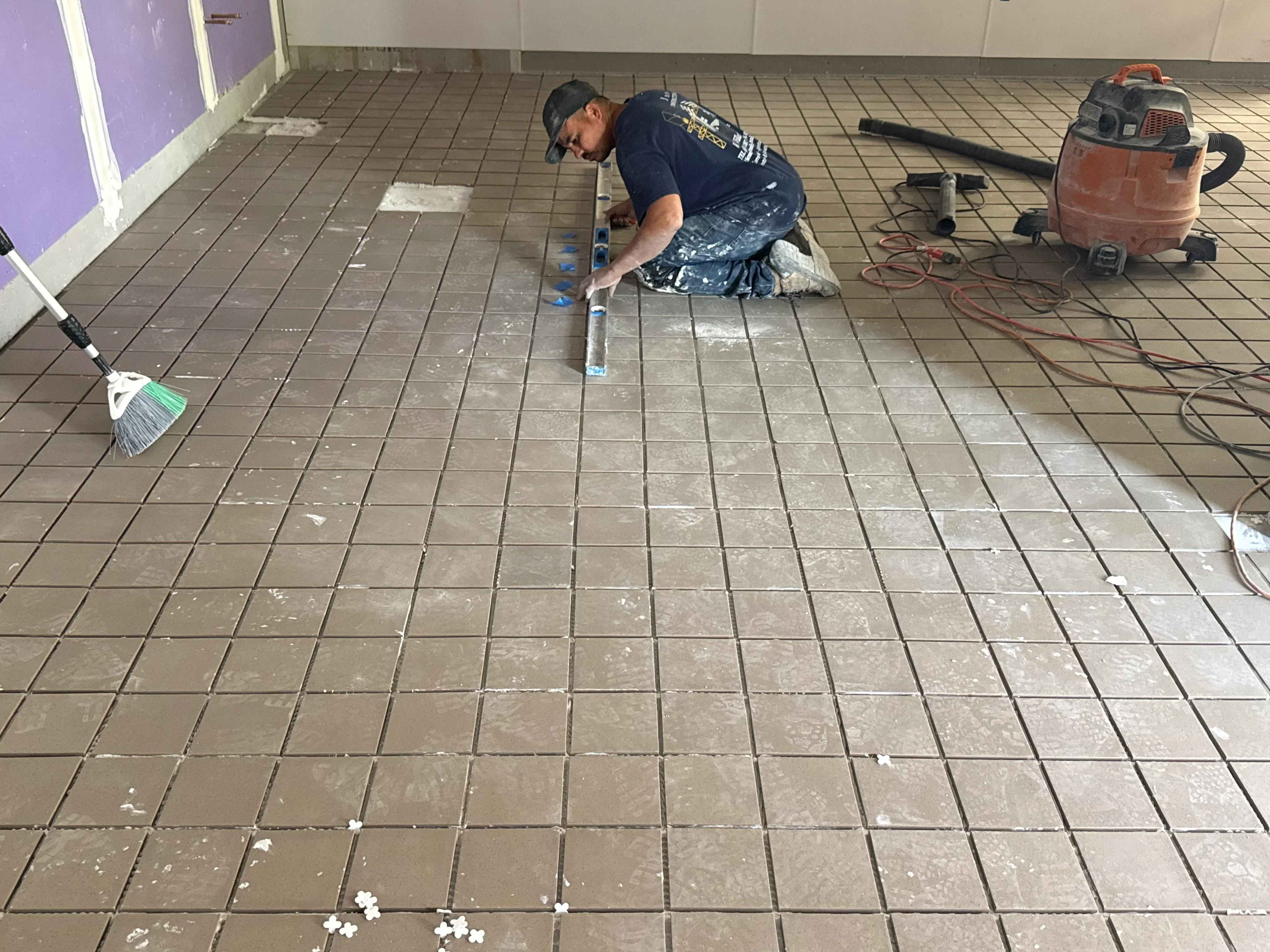 Worker installing ceramic tile flooring with grout lines and leveling spacers on commercial floor